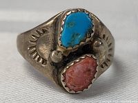 Front angle showing raw blue turquoise and red coral stones set side by side with sawtooth bezel and sunray stamp design on silver band.