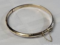 Full view of circular sterling silver hinged bangle bracelet with carved decoration and safety chain.