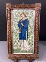 Full view of framed hand painted tile showing a young man playing a horn in blue and white clothing against a floral green foliage background.