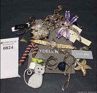 Photo of seven-piece set including Lydell NYC bracelet, peacock and dragonfly brooches, candy cane, beaded cat key chain, pig key chain with Cara NY tag, and gold-tone starfish letter 'J' key chain on dark background.