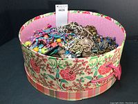 Hatbox opened showing a large quantity of mixed costume jewelry in a floral patterned hatbox, items include beaded necklaces, chains and other jewelry pieces in various colors and materials.