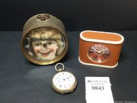 Three clocks displayed together: antique chatterbox with smiling girl face, pocket watch, and Westclox plastic travel alarm clock