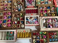 Overhead view of multiple open cardboard boxes filled with assorted round and elongated glass ornaments in pink, blue, gold and silver.