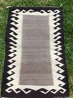 Top down photo showing full view of handmade Native American pattern rug laid flat on grass. Shows rectangular shape with gray center and geometric black and cream border.