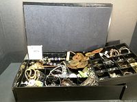 Open large black plastic jewelry box showing compartments filled with assorted costume jewelry including necklaces, bracelets, and earrings.