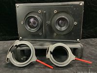 Front view of black metal enlarger mount plate holding two lenses and two red-handled adjustment arms in front.