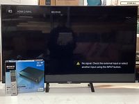 Sony Bravia TV displaying 'No signal' message, Blu-ray DVD player box and Blu-ray remote beside it
