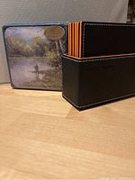 Photo showing unopened Carpentree coasters featuring a fishing scene in packaging, and a set of black and orange faux leather coasters in a storage box by Magic Vosom.