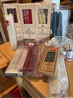 Photo showing a variety of candle boxes and loose candles including white, cream, red, burgundy, and black tapers, some in original packaging, alongside tealight candles in different packaging.