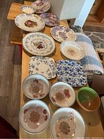Overall assortment of plates and bowls laid out on wooden bench and floor, showcasing various floral patterns including square and round plates and some stoneware bowls