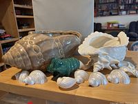 Two ceramic shell planters, one light brown and one off-white, with a green ceramic caterpillar watering accessory and eight pearlized real shell decor pieces on a table.