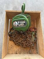 Green metal 1 ton chain hoist with attached steel chain and hook inside a wooden box.