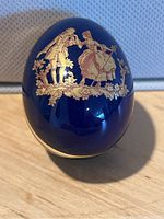 Closed view of the blue porcelain egg with detailed gold pastoral scene of a man and woman in period dress