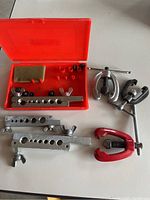 Photo of assorted flaring tools including bar clamp flaring tools, yoke flaring tools, a red C-clamp tool, and a red box containing tube adapters and a sponge