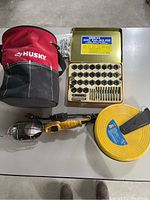 Lot overview showing the Husky tool bag, 60-piece Tap and Die set in metal tin, retractable trouble light and cable reel