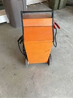 Full view of the orange metal industrial battery charger on wheels with handle and attached cables with clamps.