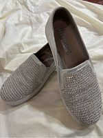 Pair of silver slip-on Skechers shoes with rhinestones and pearl accents displayed on white fabric background.