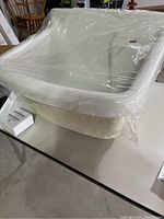White laundry sink basin wrapped in clear protective plastic film, showing front and side, placed on table.