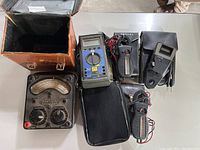 Seven ammeters displayed with vintage analog meter, digital meters, clamp meters, and carrying cases on table.