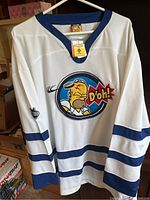 White hockey jersey with blue collar and stripes on sleeves, Homer Simpson embroidered logo with 'D'oh!' comic bubble in center front, showing new condition with tags attached.