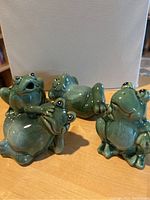Front view showing all four ceramic frog figurines grouped on a wooden surface against a white backdrop.