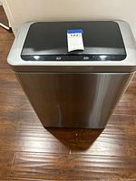Frontal view of the stainless steel automatic trash can with black sensor lid, placed on a wooden floor.