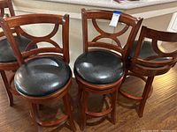 Four wooden swivel barstools with black leather seats arranged on hardwood floor.