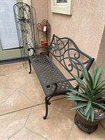Photo showing the black metal outdoor bench with decorative scrollwork and lattice seat, positioned next to a large potted plant.