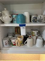 White ceramic teapot and various coffee mugs with different designs on a shelf.