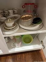 Shelving unit displaying various white ceramic dishes and mugs with floral designs, glass bowls, and plastic containers.