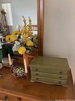 Green jewelry box with three drawers, floral arrangement, grapevine candle holders with candles, and a drawer visible in mirror