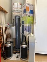 Photo showing two black and silver air cleaners, one tall oscillating tower fan in front of the boxes labeled 2 FANS, another box fan partially visible behind, and white baby gates leaning against the wall.