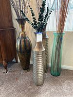 Four floor vases with different finishes and decorative sticks, placed on carpet near furniture and window light.