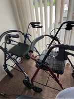 Photo showing four walkers grouped together by a window; maroon rollator with basket, blue rollator with padded seat, and two additional walkers including one with a black cushion.