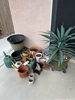 Overview of pots, plant, watering cans, and other gardening tools on concrete surface against house wall.