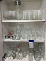 Shelves displaying glassware including short glasses, crystal wine glasses, beer mugs, shot glasses, and glass mugs arranged in multiple rows.