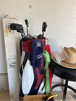 Golf bag with golf clubs inside, headcovers visible, also red cooler jug and black barstool with straw sun hat on it.