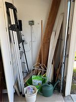 Full view of ladder, gardening tools including watering can, broom, wooden planks and more.
