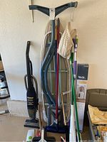 Photo of ironing boards, Hoover QuickBroom vacuum, various brooms, mops, plunger, and hangers arranged in garage against wall.