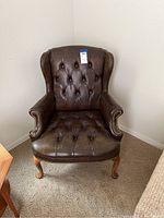 Front view of dark brown leather wingback chair with button tufting, brass tacks along armrests, and ornate wooden legs.