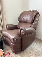 Brown leather recliner chair positioned in corner of room showing side and front view. Upholstered in smooth brown leather with brass tacks accents.