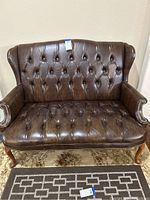 Front view of brown leather loveseat showing diamond-tufted upholstery and wooden Queen Anne legs on floral rug.