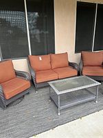 View of one loveseat, two glider chairs with orange cushions, glass-top table and grey woven rug scattered with leaves.