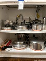Photo showing multiple stainless steel pots and pans with copper bottoms and a tea kettle placed inside a white shelving unit.
