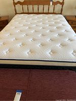 Full view of the queen size mattress with textured quilting and navy blue piping on the sides, sitting on a dark boxspring and partially covered by a brown bed ruffle; behind is the maple wooden headboard with spindle design visible.
