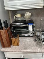 Kitchen countertop showing Oster stainless steel toaster oven, two wooden knife blocks with multiple knives, and assorted coffee carafes and coffee pot