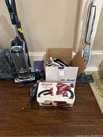 Full view of Shark upright vacuum, Shark floor steam cleaner, Dirt Devil hand vacuum in box, and box of hangers on floor against wall.
