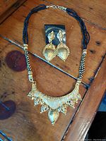 Full view of the gold-tone textured pendant necklace with braided black bead strands paired with matching leaf-shaped earrings.