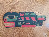 Top view of carved and painted wooden wall art with Whale & Raven design in green, red, black and beige colors, showing surface details on hardwood floor.