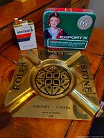 Photo showing the brass advertising ashtray with Rolph Stone Limited branding, Vantage cigarette lighter, and Export A metal container